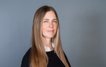 Professional headshot of a woman with long light brown hair wearing a black sweater over a white collared shirt, standing against a gray background.