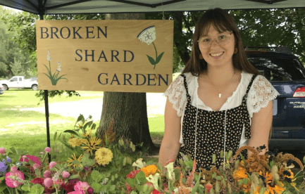 A woman with brown hair in black overalls, standing under a tent, a table with a floral table cloth and bouquets of flowers in mason jars in front of her. A sign behind her reads Broken Shard Garden.