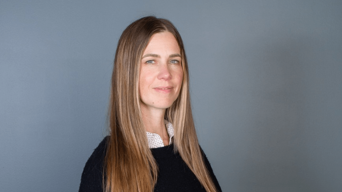 Professional headshot of a woman with long light brown hair wearing a black sweater over a white collared shirt, standing against a gray background.