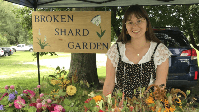 A woman with brown hair in black overalls, standing under a tent, a table with a floral table cloth and bouquets of flowers in mason jars in front of her. A sign behind her reads Broken Shard Garden.